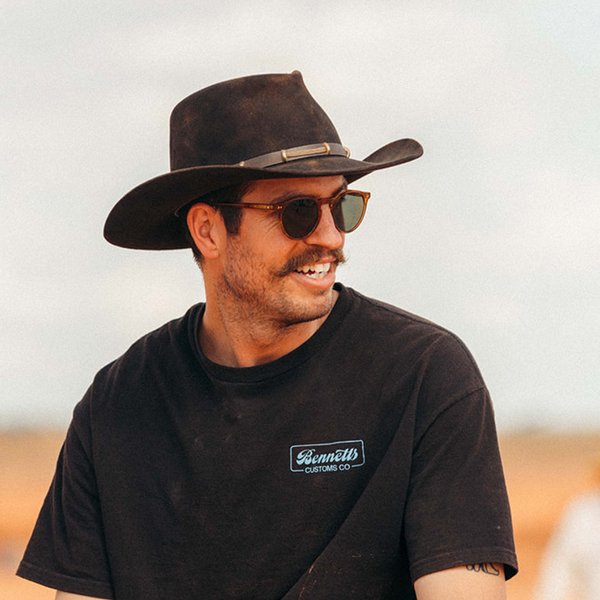 Man wearing round sunglasses, a cowboy hat and a black shirt.