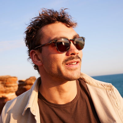 Man wearing scratch resistant sunglasses in front of red rocks and the ocean