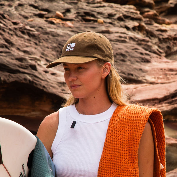 Surfer girl standing with an army green cap on and holding her surfboard
