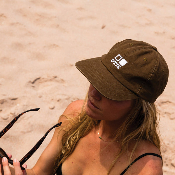 Woman wearing army green cap holding OTIS eyewear sunglasses