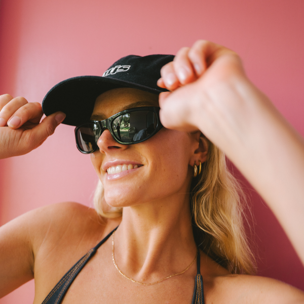 Blone woman smiling wearing black cap, black bikini and black wrap sunglasses