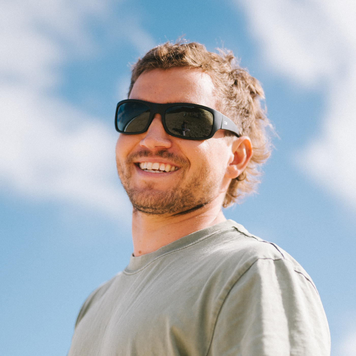 A smiling man wearing black sunglasses and a light olive green t-shirt, photographed outdoors against a bright blue sky with soft white clouds.