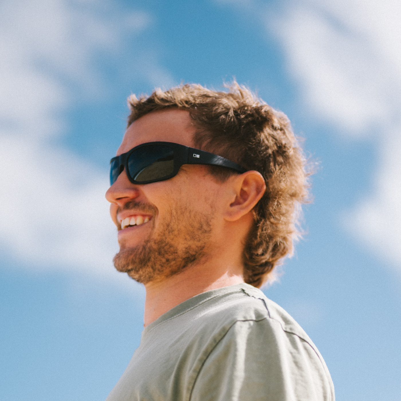 A man in side profile wearing black sunglasses with an OTIS logo and a light olive green t-shirt, set against a bright blue sky with soft white clouds.