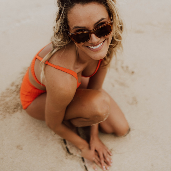 Woman wearing orange bikini and sunglasses sitting on the sand and smiling