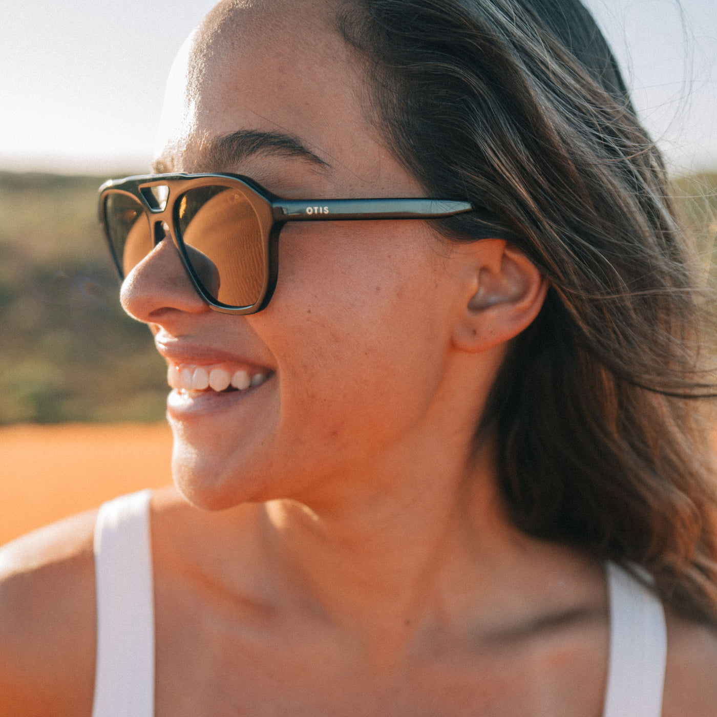 A close-up lifestyle shot of a woman wearing OTIS sunglasses outdoors. The sunglasses feature a thick black frame, warm brown mirrored lenses, and a distinctive teal-blue temple arm with the brand name OTIS printed in white.
