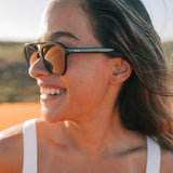 A close-up lifestyle shot of a woman wearing OTIS sunglasses outdoors. The sunglasses feature a thick black frame, warm brown mirrored lenses, and a distinctive teal-blue temple arm with the brand name OTIS printed in white.