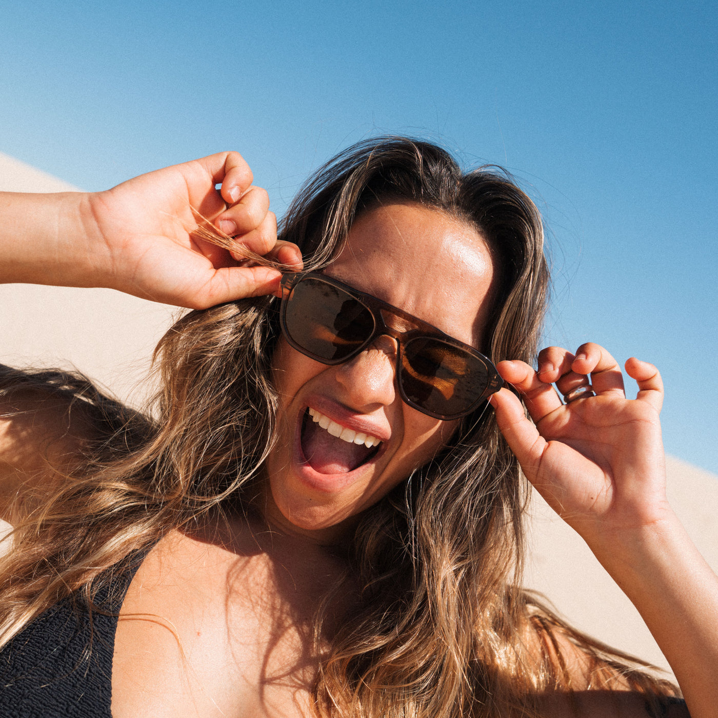A woman outdoors wearing dark brown aviator-style sunglasses, holding them in place with both hands against a bright blue sky and sandy background.