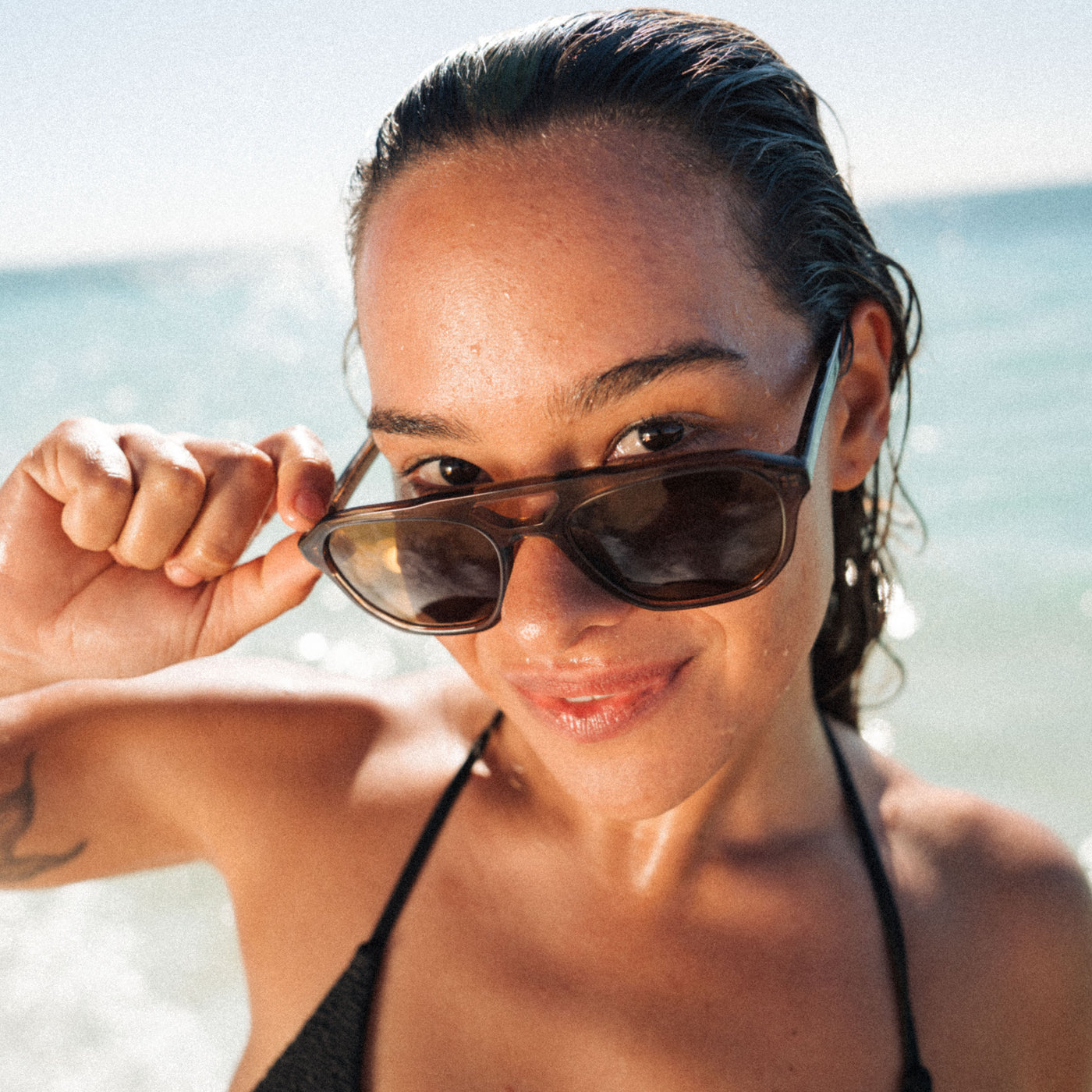 A woman at the beach wearing dark brown aviator sunglasses with a thick black frame, holding them slightly off her face with one hand.