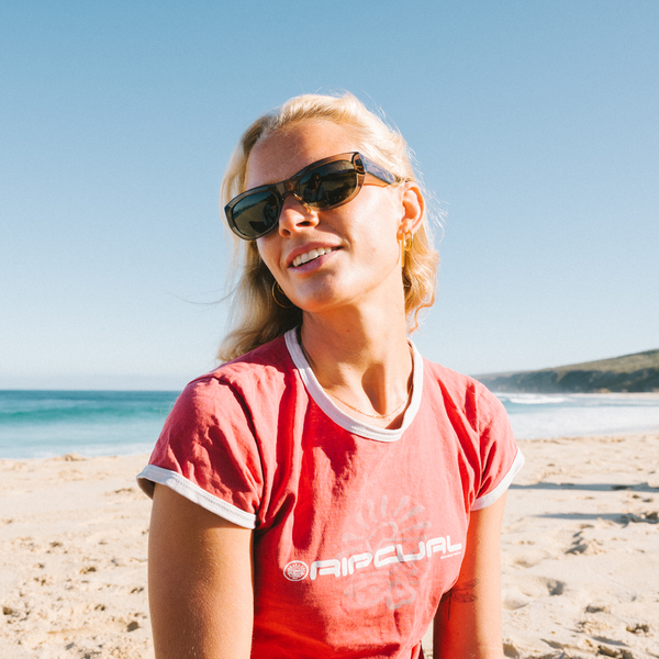 Woman wearing sunglasses and a red shirt with a ripcurl logo on a beach