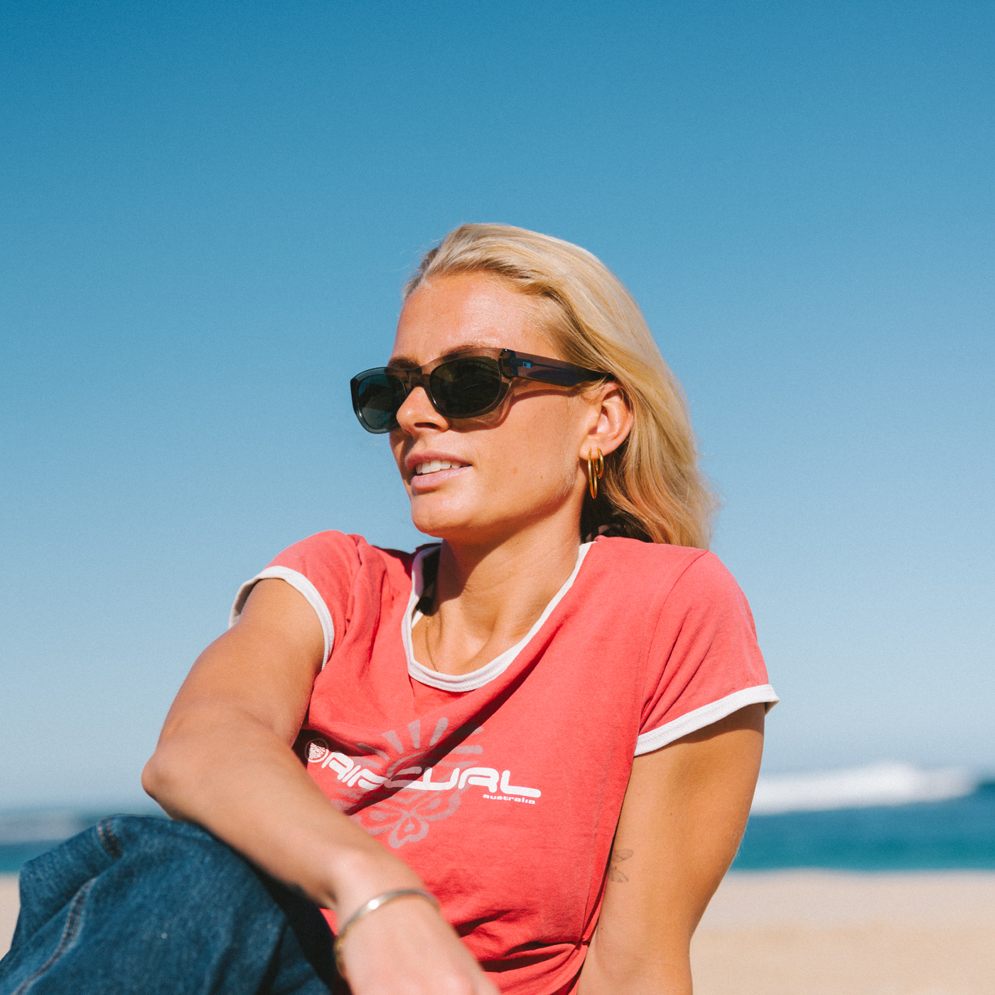 Woman wearing sunglasses and a red shirt sitting on a beach with clear blue sky.