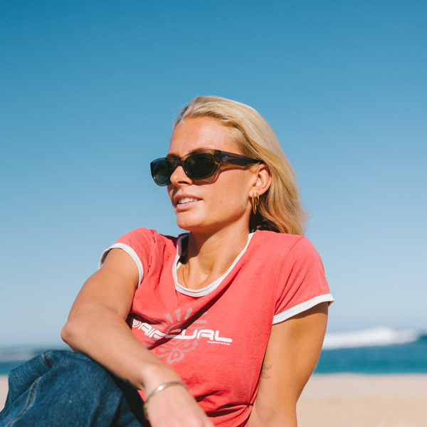 Woman wearing sunglasses and a red shirt sitting on a beach with clear blue sky.