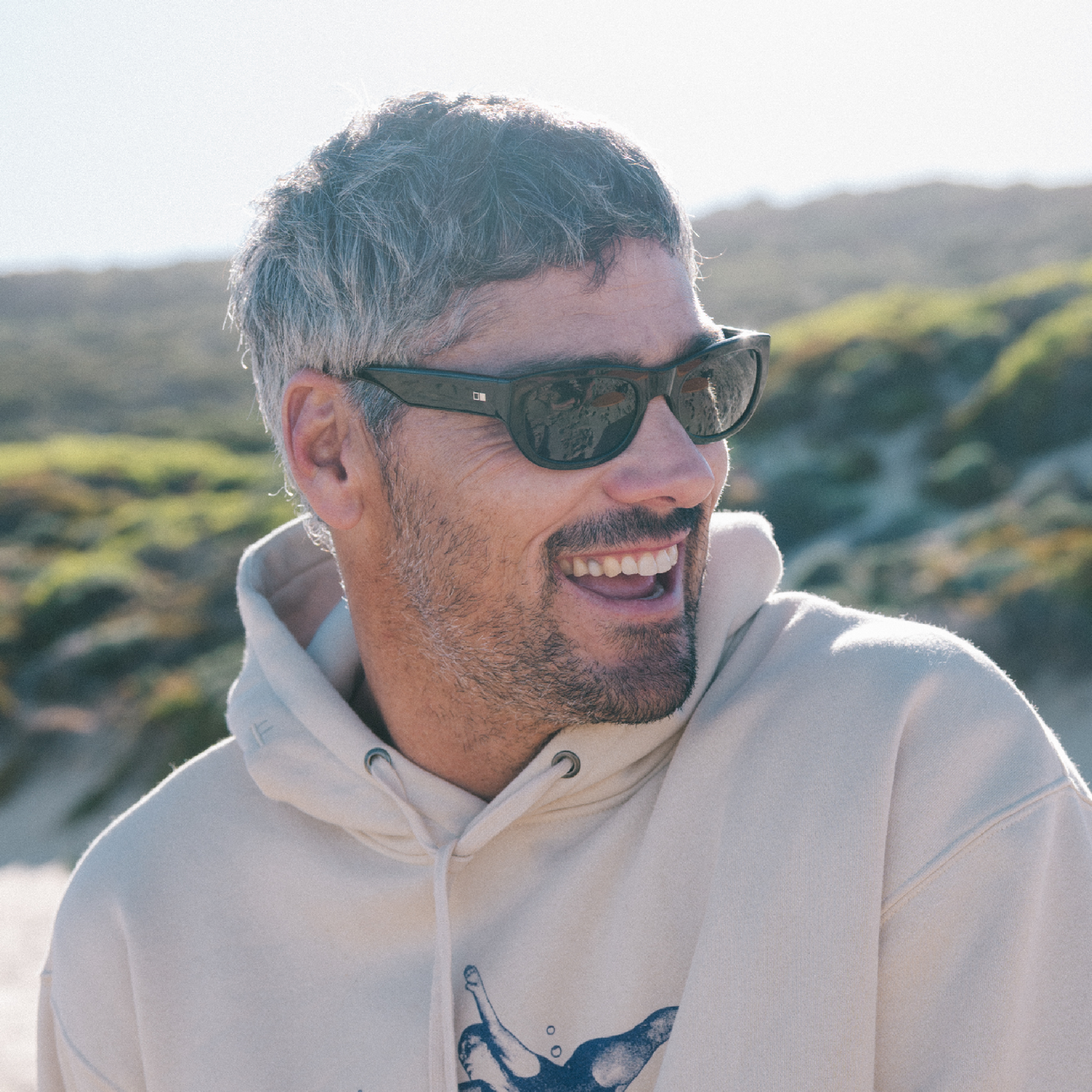 Man wearing sunglasses and a beige hoodie with a scenic background