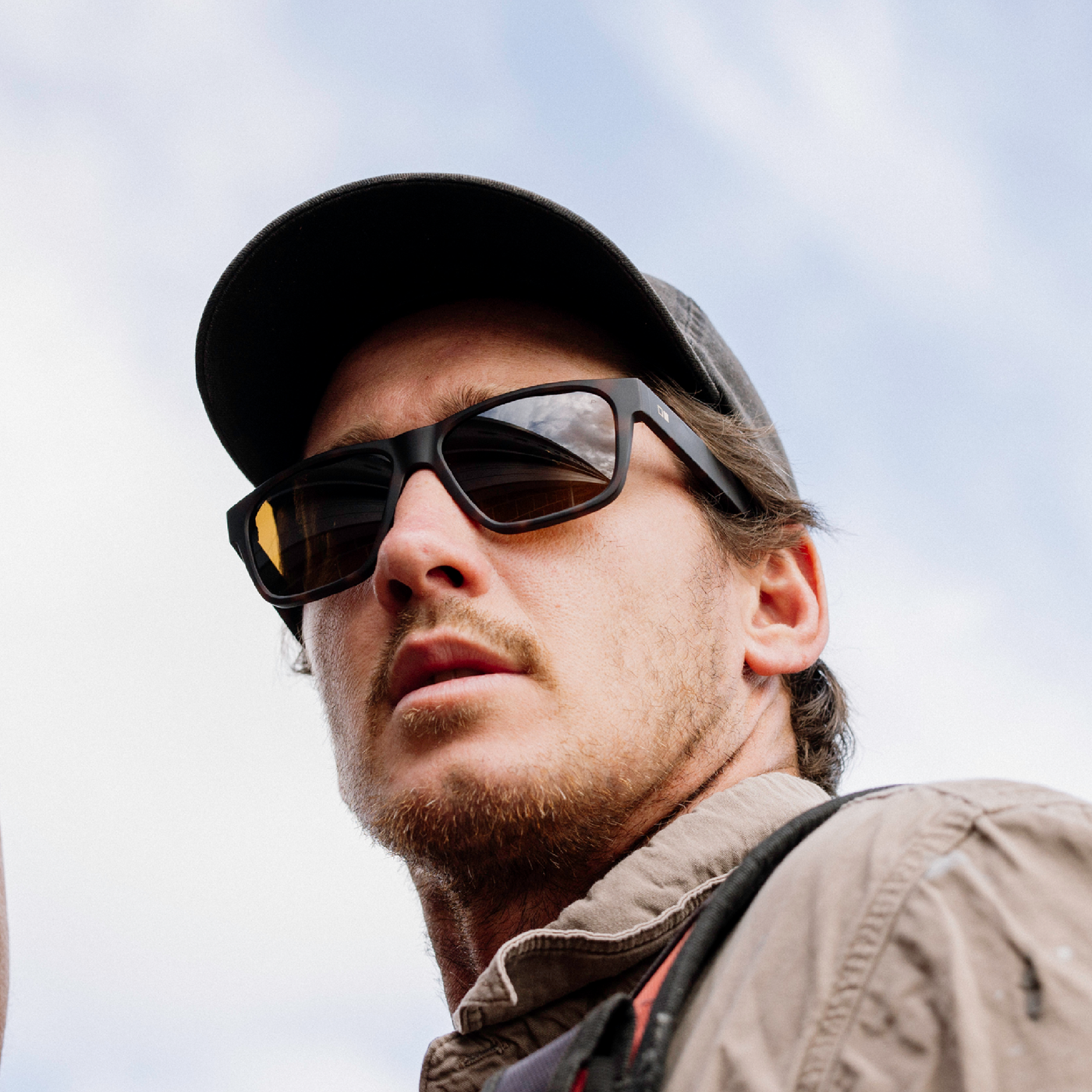 A man wearing black rectangular sunglasses with a black baseball cap and a beige jacket, photographed outdoors against a cloudy sky.