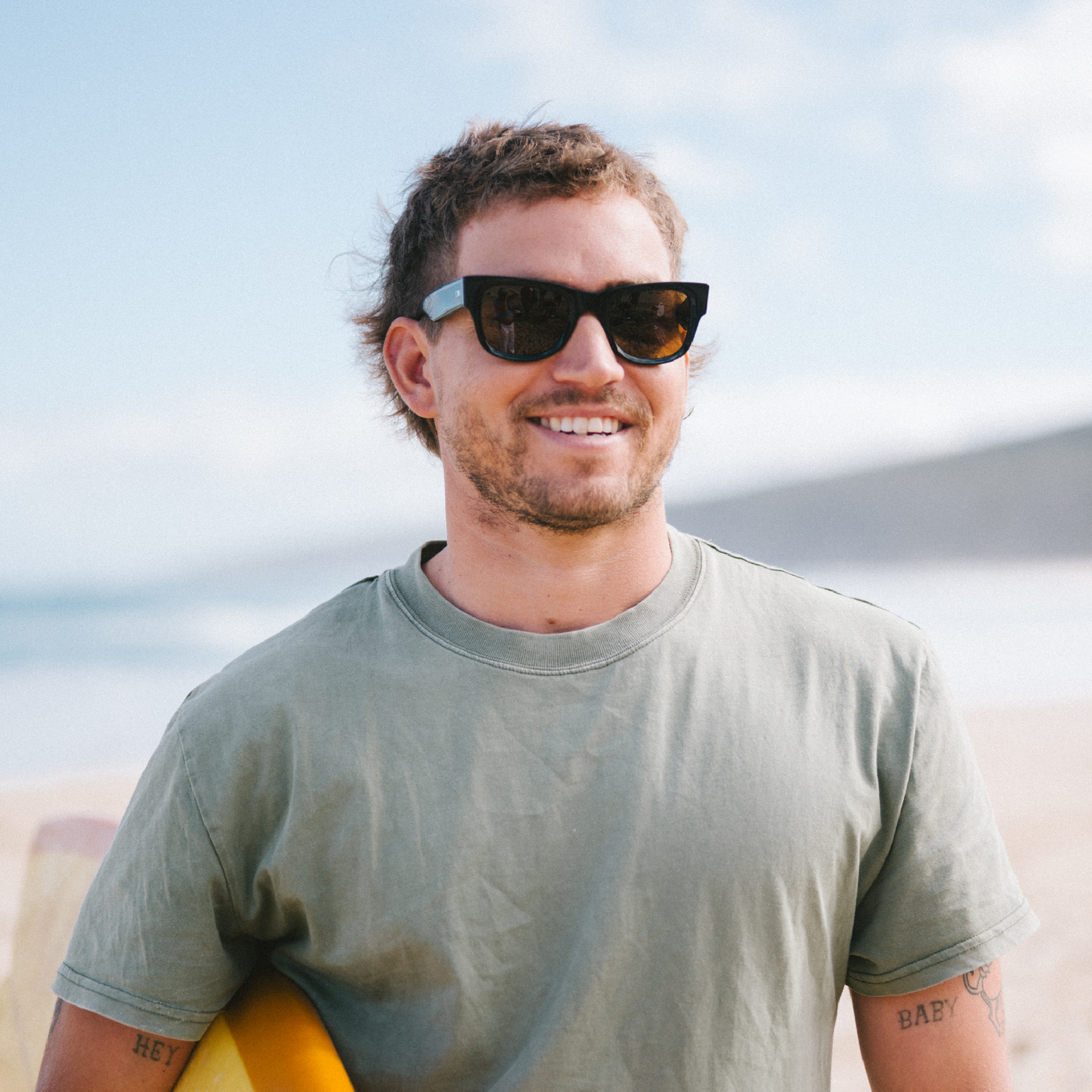 Man wearing wayfarer sunglasses and a green t-shirt holding a surfboard on a beach.
