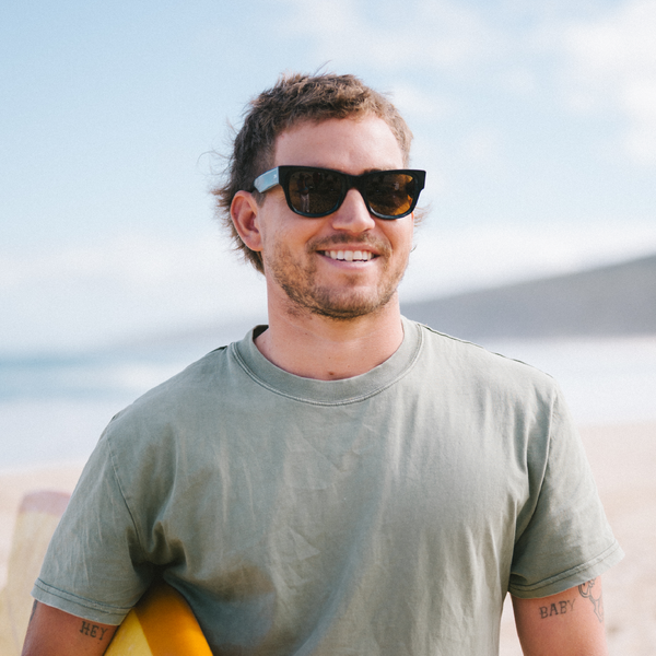 Man wearing wayfarer sunglasses and a green t-shirt holding a surfboard on a beach.