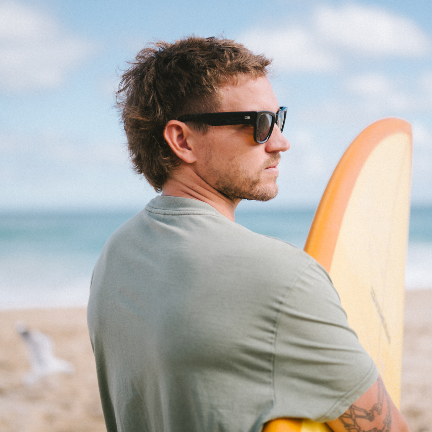 Man wearing wayfarer sunglasses and a green t-shirt holding a surfboard on a beach.