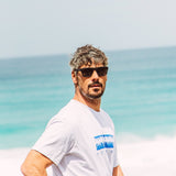 Jay Davies looking at the camera in black sunglasses wearing a blue and white t shirt in front of a blue ocean background