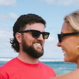 Two people laughing and wearing sunglasses with a beach and ocean background