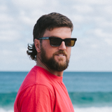 Man wearing square sunglasses and a red shirt with ocean and sky in the background