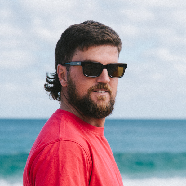 Man wearing square sunglasses and a red shirt with ocean and sky in the background