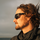 A man in profile wearing black sunglasses with a visible OTIS logo on the temple, set against a soft, overcast sky background.