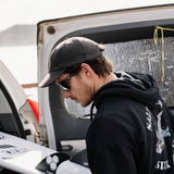 A man wearing a black baseball cap, sunglasses, and a black hoodie leans near an open white vehicle door in an outdoor setting.