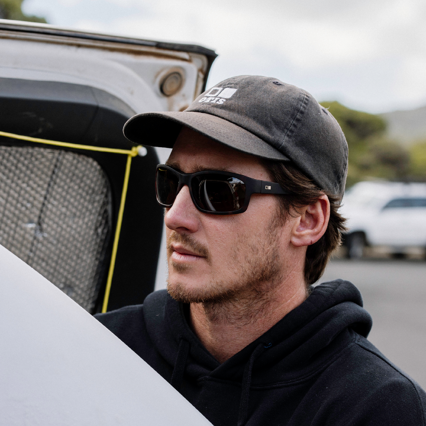A man wearing a dark gray baseball cap with a white logo, black sunglasses, and a black hoodie, standing outdoors next to a white vehicle.