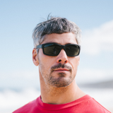 A close-up portrait of a man wearing black sunglasses and a red shirt against a bright sky background.