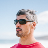 A man with gray hair and a beard wearing black sunglasses and a red t-shirt, photographed outdoors against a bright blue sky.
