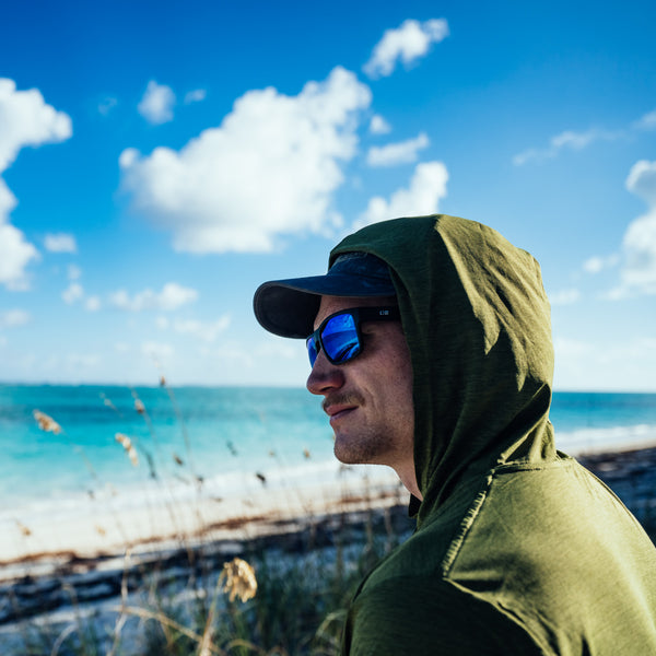 Man in green hoodie and blue reflect sunglasses and hat sitting on beach