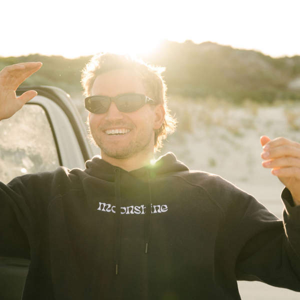 Man wearing wrap sunglasses and a black hoodie with 'moonskies' text, outdoors with sunflare.