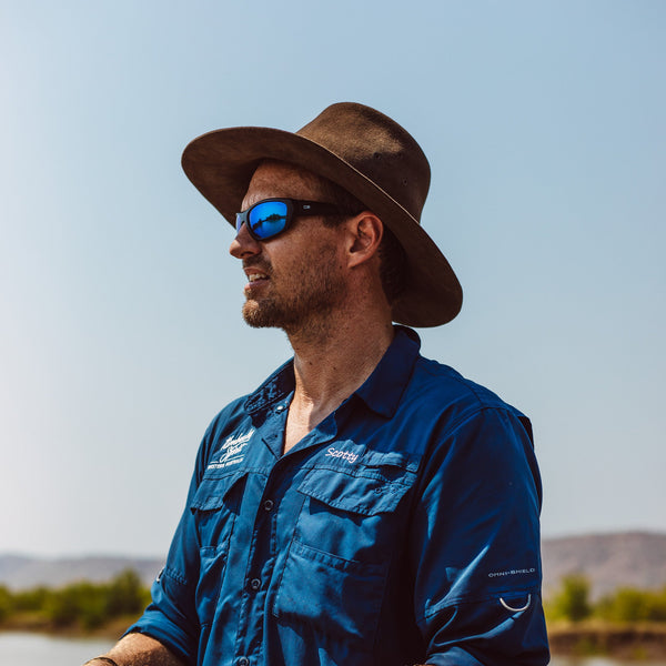 Man wearing a blue shirt and brown hat with sunglasses, standing by a body of water.
