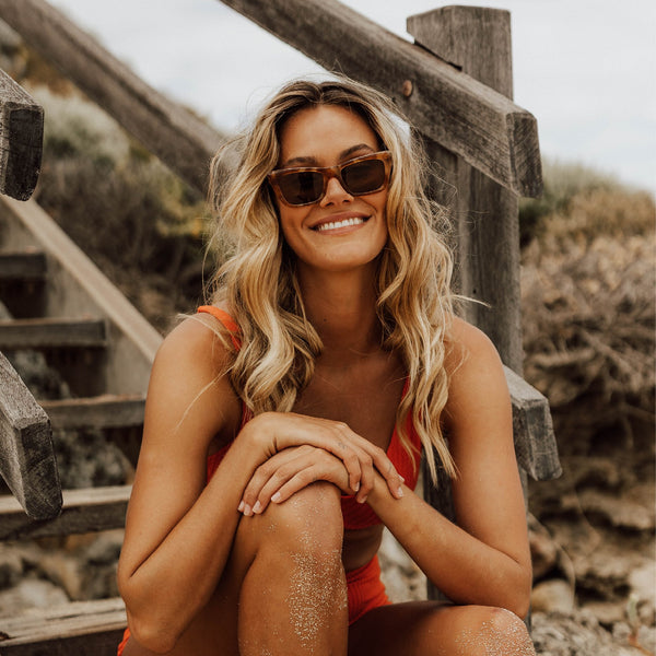 Blonde woman sitting on wooden stairs smiling wearing tortoise shell sunglasses