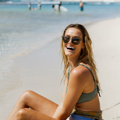 Girl sitting on beach laughing and wearing crystal blue sunglasses