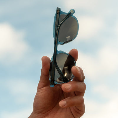 Hand holding blue sunglasses in the sky in front of the clouds