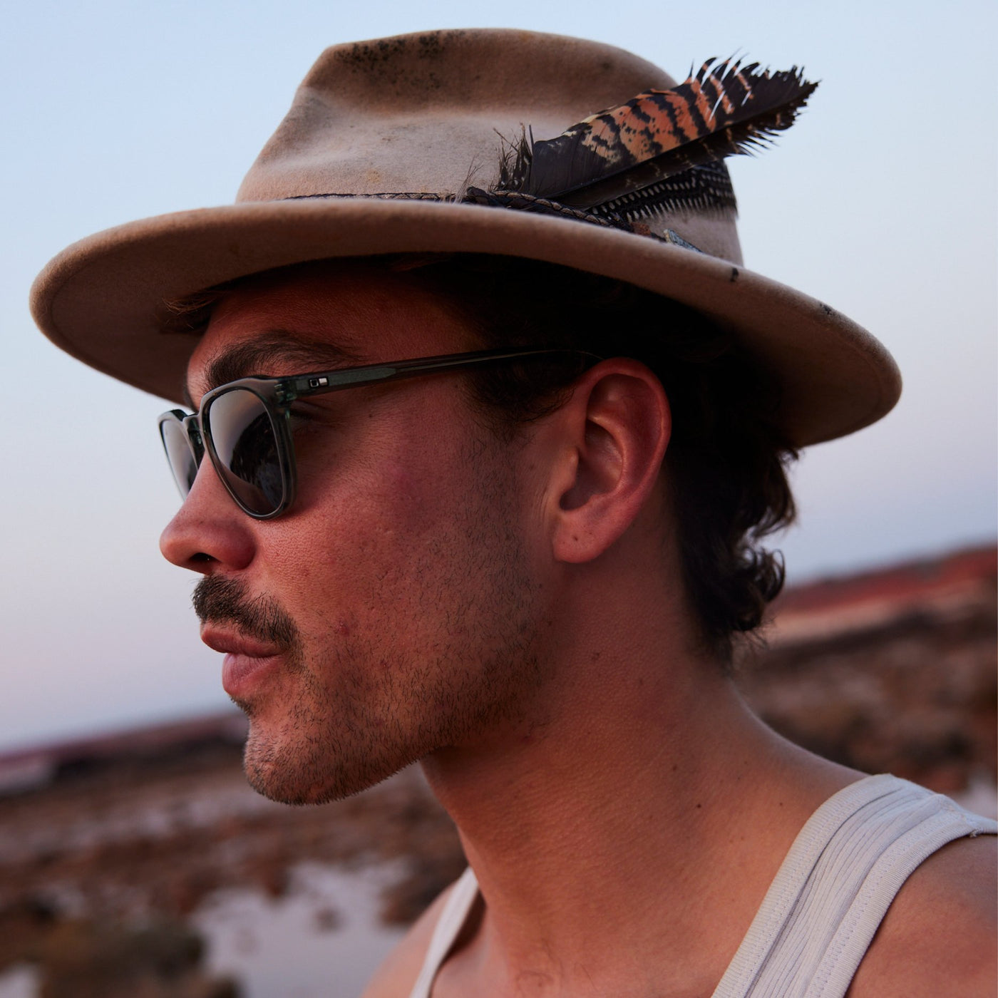 Man wearing Akubra hat with a feather in it and blue sunglasses looking out to the distance