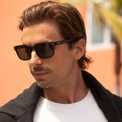 Man wearing wayfarer sunglasses with a white top and black jacket over his shoulders