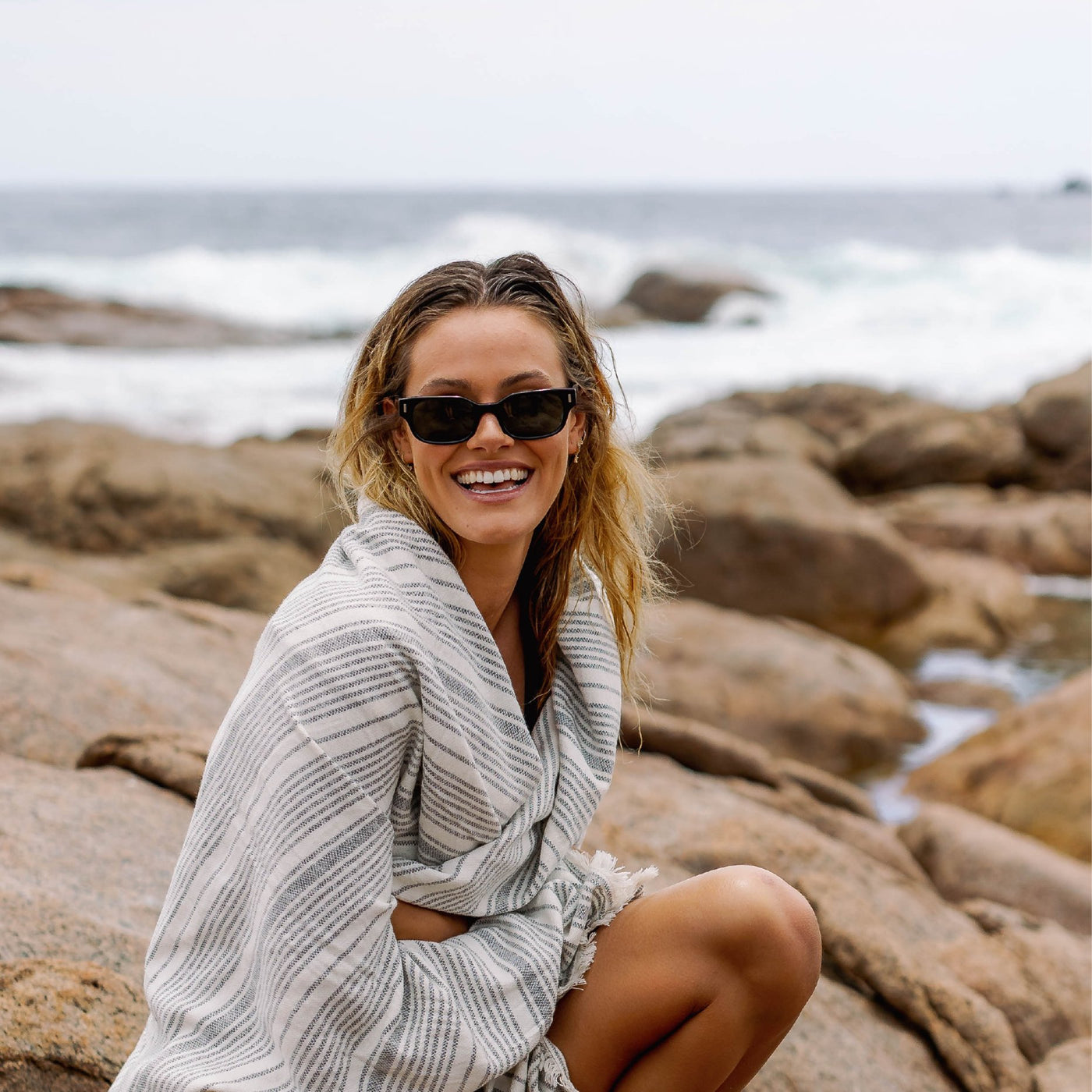 Girl sitting on a rock by the beach wearing a towel wrapped around her and black sunglasses