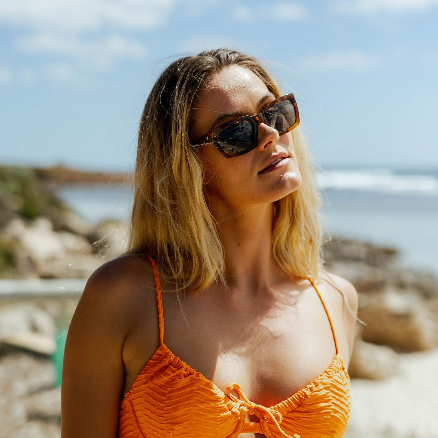 Woman wearing rectangle sunglasses and an orange bikini top at the beach
